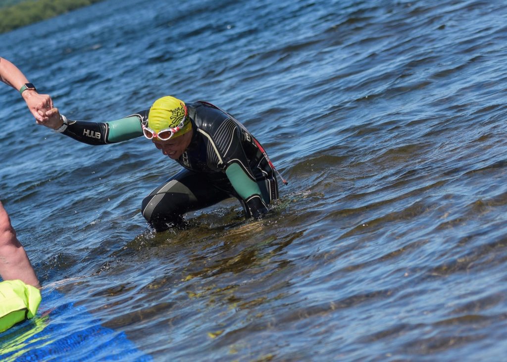 A swimmer in a wetsuit being given a helping hand to climb out of a lake