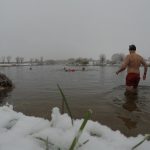 A man standing knee deep in water facing away from the camera. Other swimmers in the distance. Snow on the ground in the foreground.