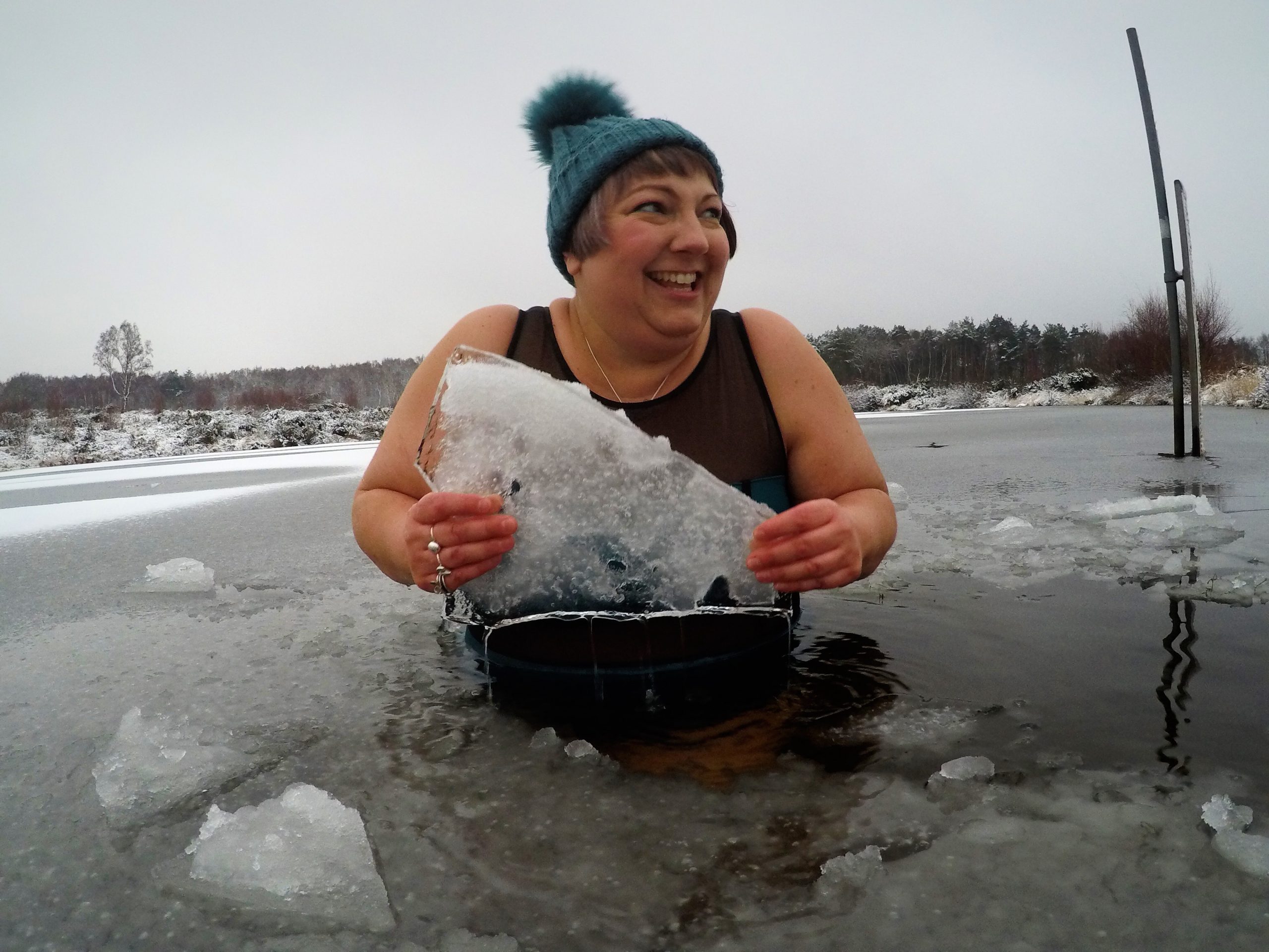 Female standing in cold water holding block of ice. wearing a swimming costume and wooly hat.