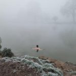A woman standing in a river on a foggy morning, back to the camera.
