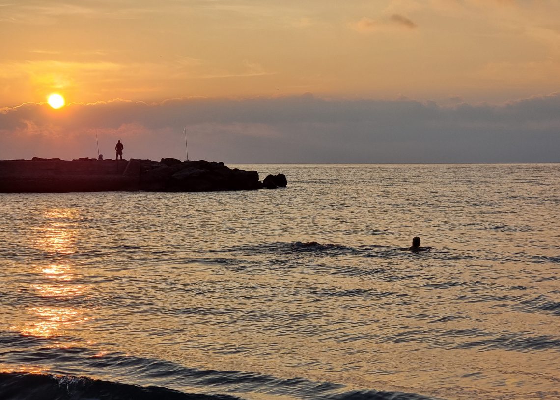 Swimmer in the sea at sunset. Person on rocks behind fishing.