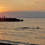 Swimmer in the sea at sunset. Person on rocks behind fishing.