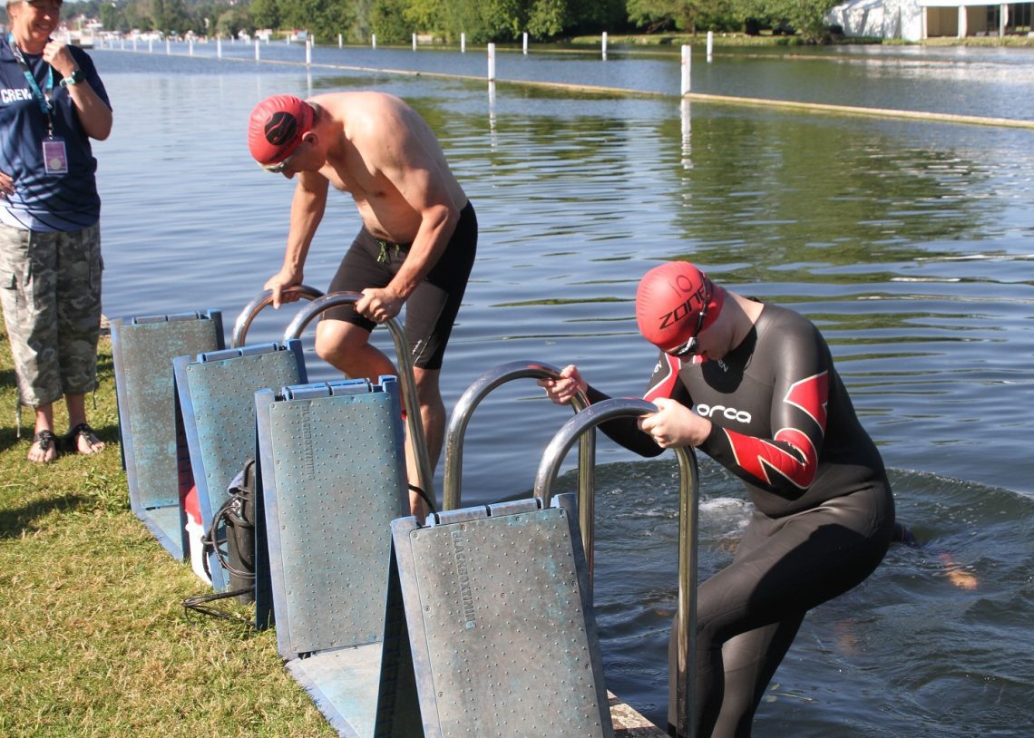 Two swimmers climbing backwards down ladders into a river. One is wearing a wetsuit.