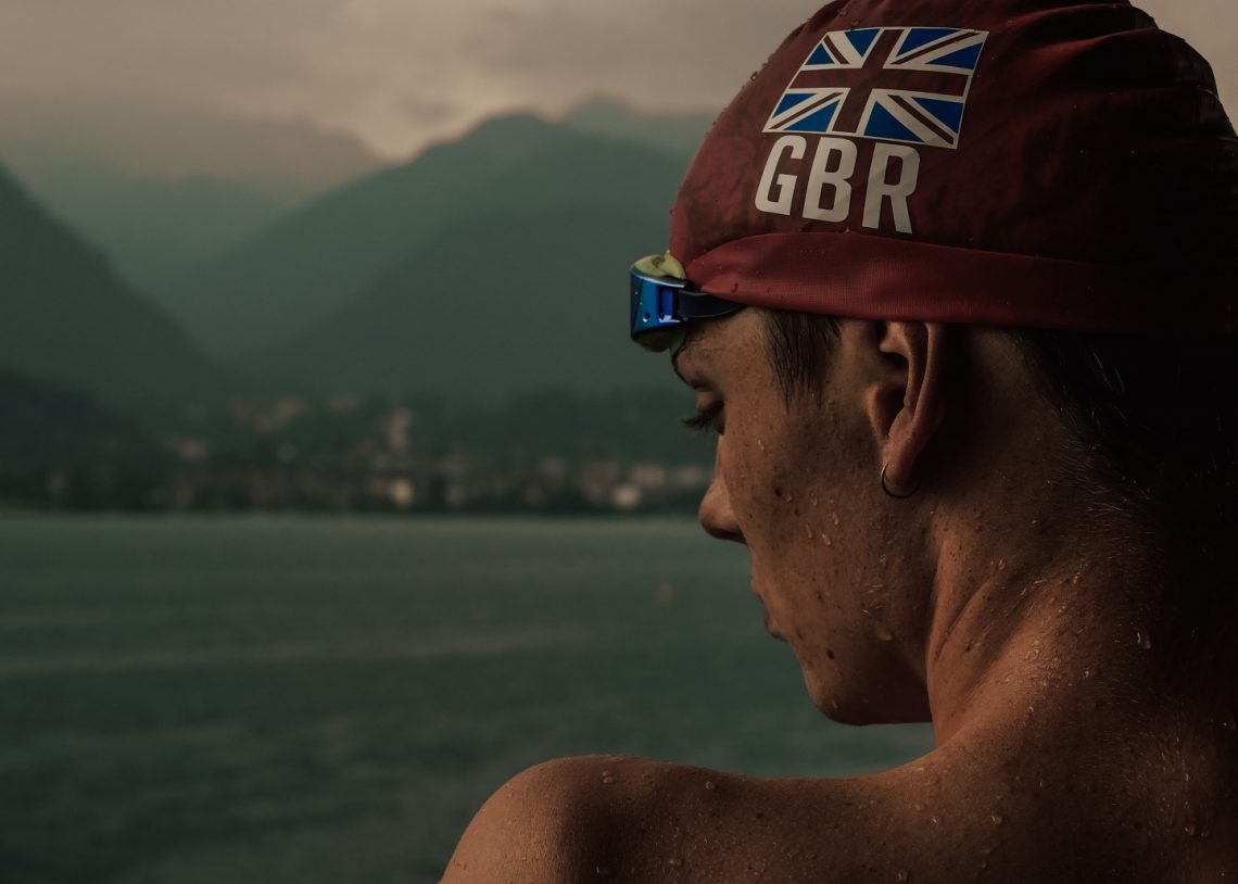 Close up of Hector Pardoe in GBR swimming cap, back to the camera, looking across a lake under stormy skies