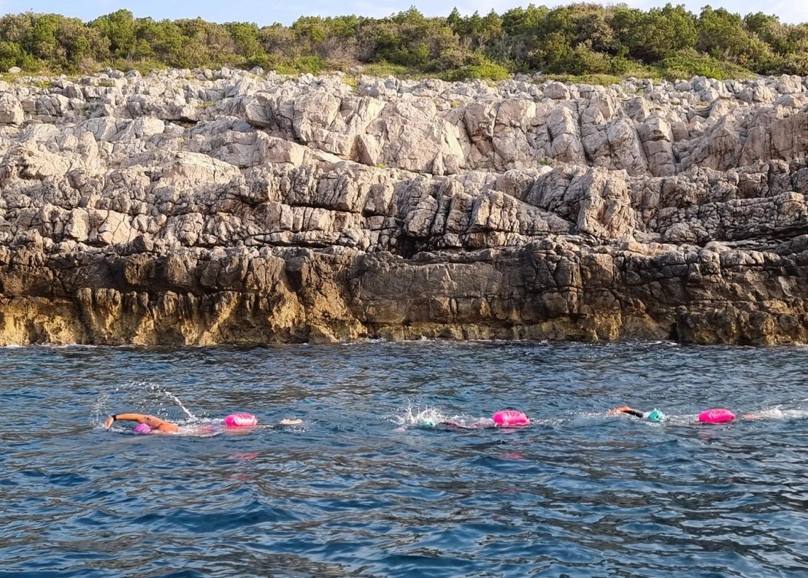 Three swimmers swimming parallel to the shore. Rocky cliffs in the background.