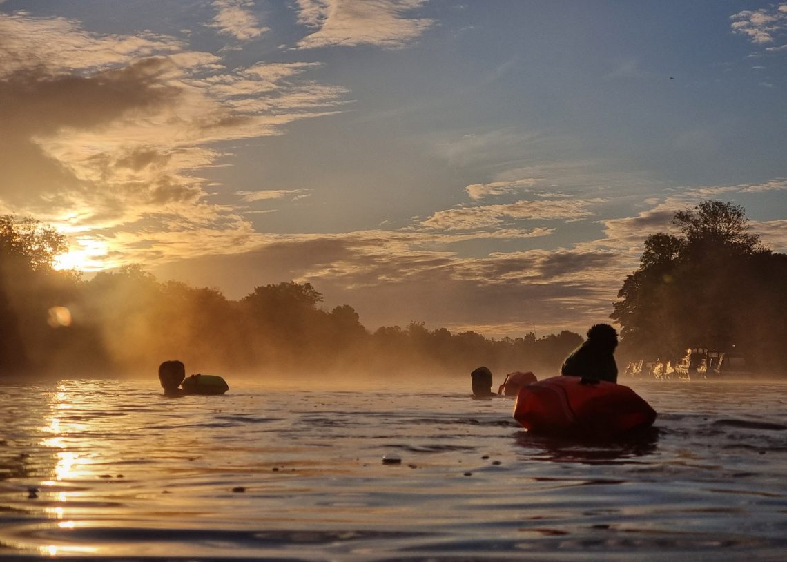 Three swimmers in silhouette in a river with the sun rising behind and to the left.